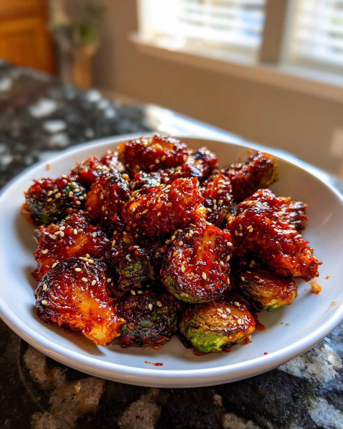 A white bowl filled with Air Fryer Crispy Gochujang Brussels Sprouts, coated in a sticky red glaze and sprinkled with sesame seeds.