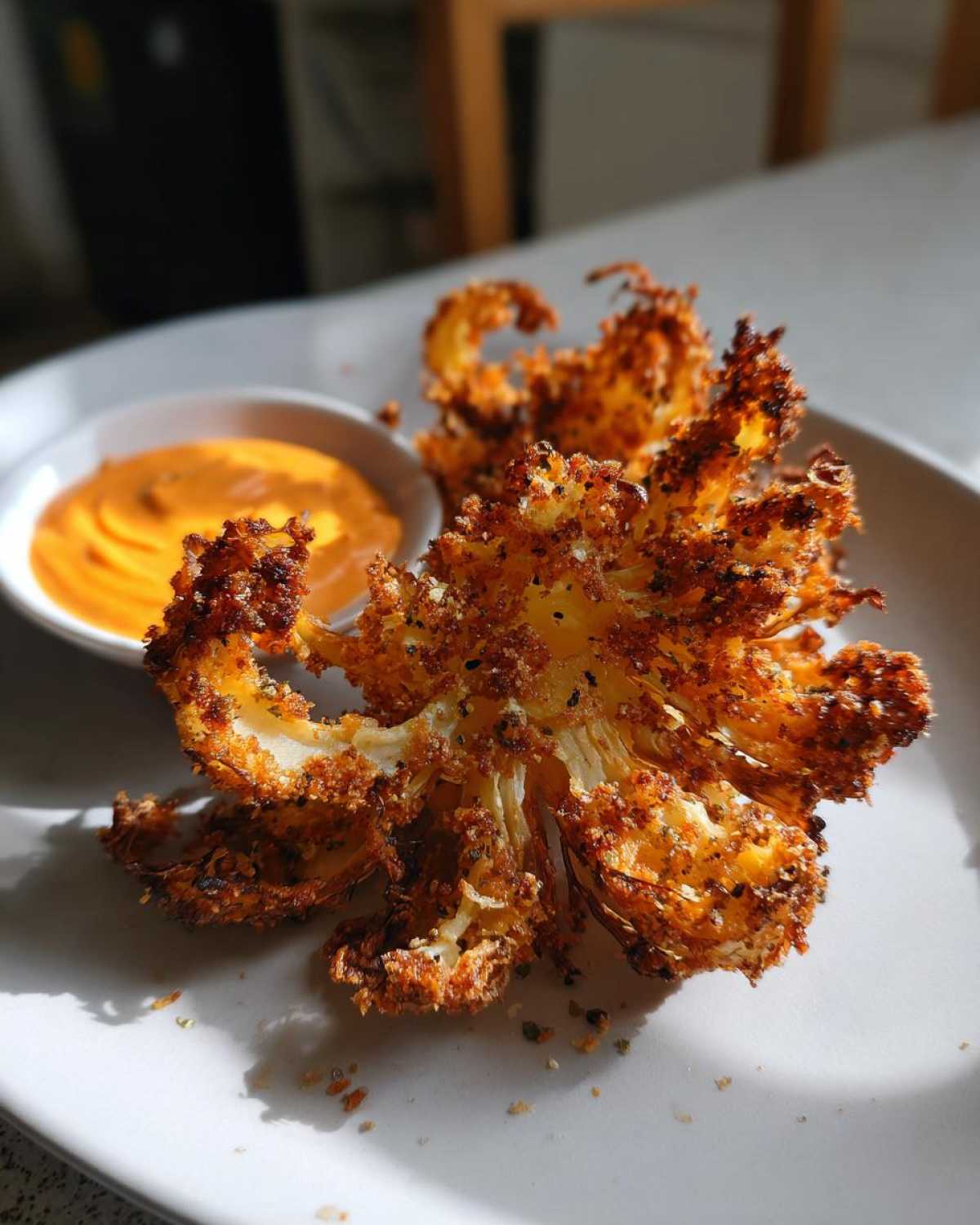 A golden brown, crispy Air Fryer Crispy Onion Petals served on a white plate next to a small bowl of spicy orange dipping sauce.