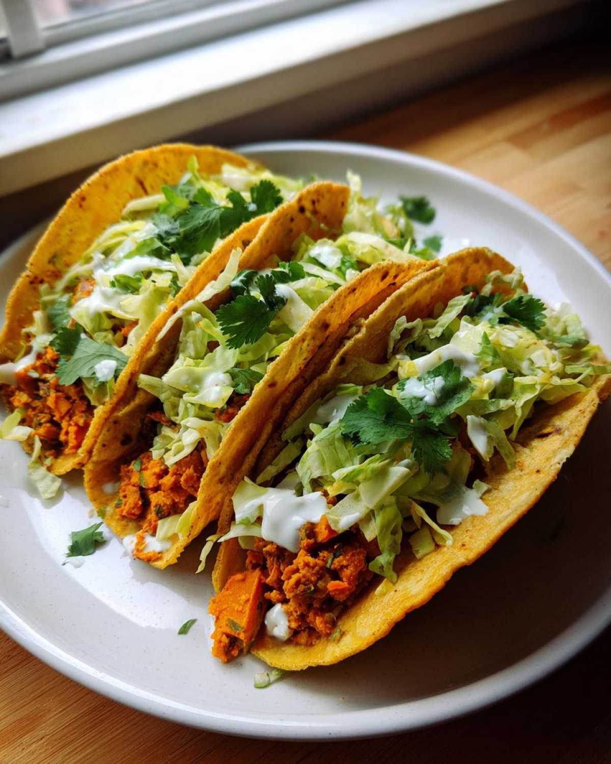 Three crispy tacos filled with orange sweet potato mixture, shredded lettuce, crema, and cilantro, ready to eat as Air Fryer Crispy Sweet Potato Tacos.