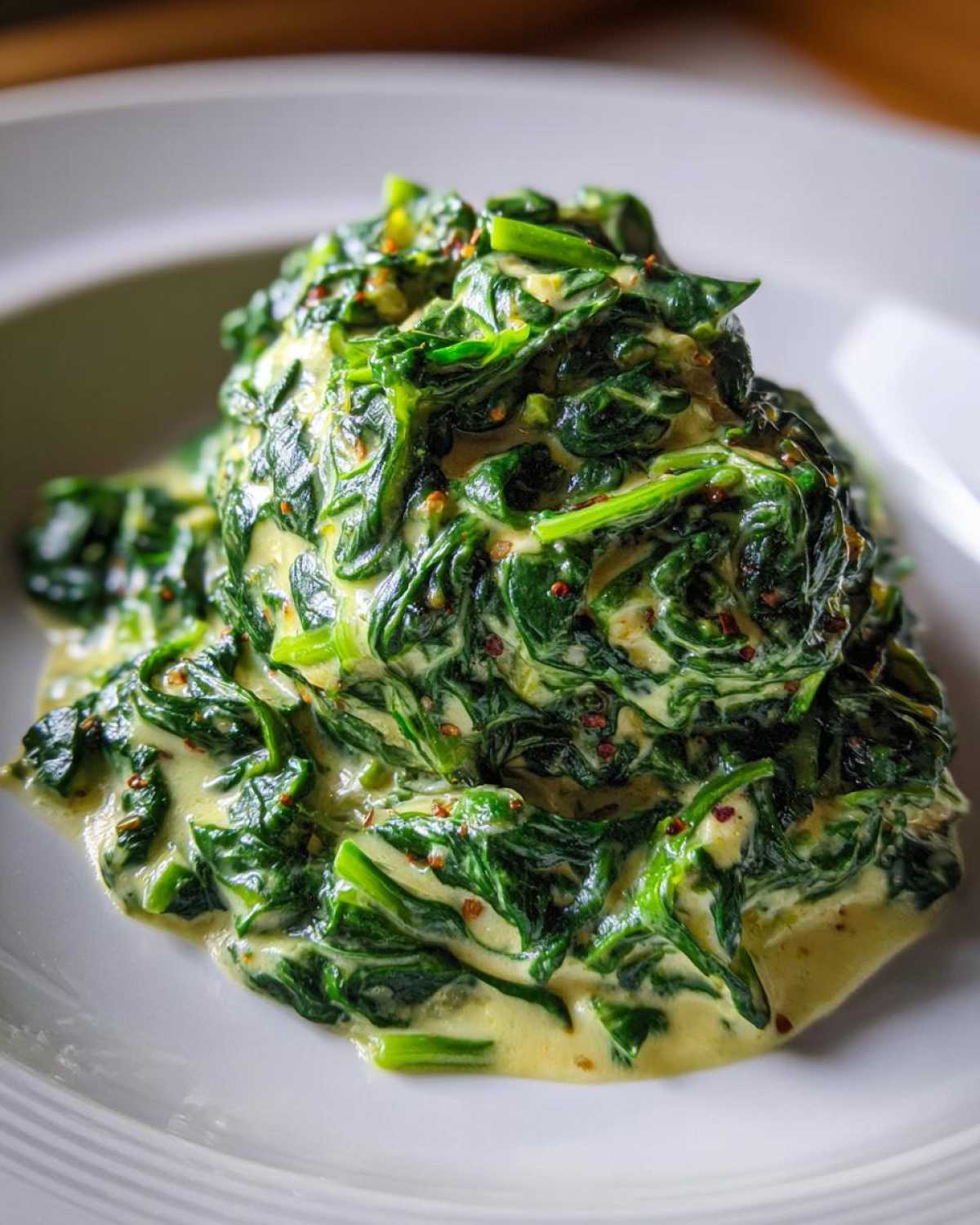 A close-up of rich, vibrant green creamed spinach served on a white plate, featuring a creamy sauce and visible spices.