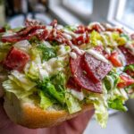 A hand holding a hearty open-faced grinder salad sandwich overflowing with lettuce, salami, and creamy dressing.