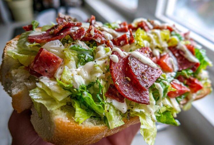 A hand holding a hearty open-faced grinder salad sandwich overflowing with lettuce, salami, and creamy dressing.