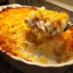 A spoonful of creamy Angel Chicken Rice Casserole lifted from a white baking dish, showing shredded chicken and melted cheddar cheese topping.