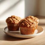Three golden brown Applesauce Oatmeal Muffins topped with oats, sitting on a light gray plate in warm sunlight.