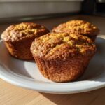 Three golden brown Applesauce Oatmeal Muffins sitting on a white plate in bright sunlight.