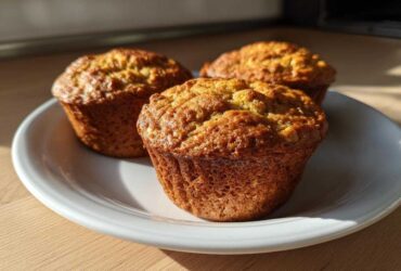 Three golden brown Applesauce Oatmeal Muffins sitting on a white plate in bright sunlight.