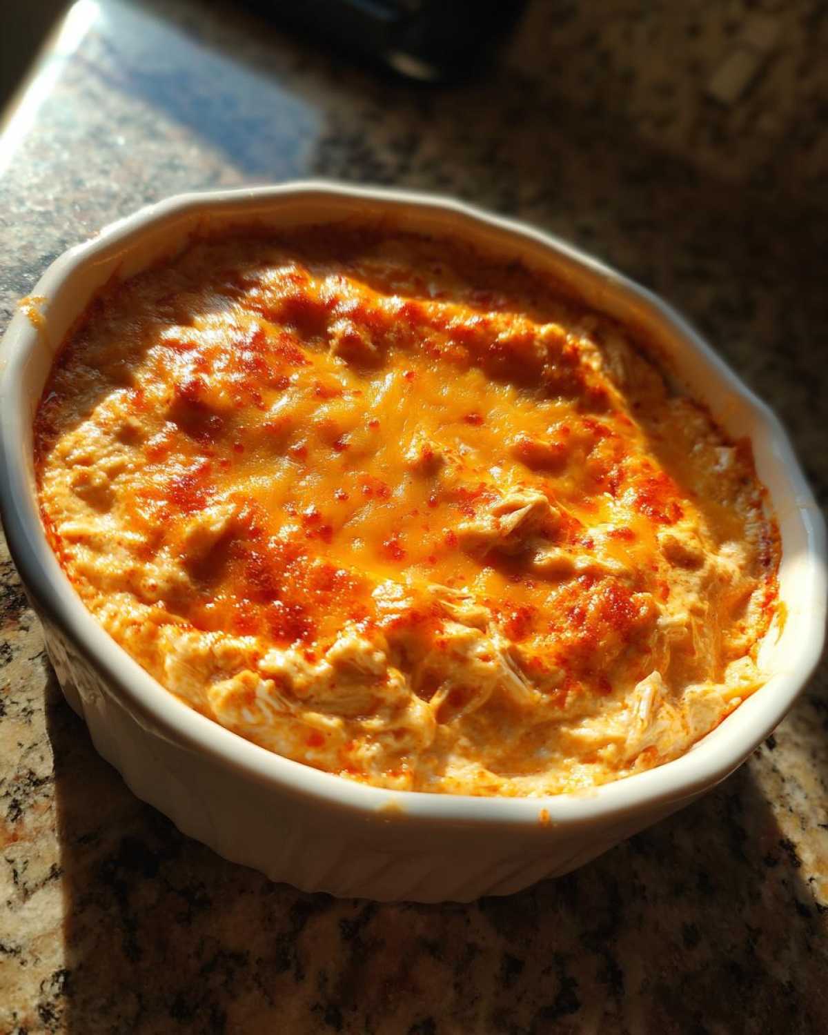 A close-up of a creamy, baked cottage cheese buffalo chicken dip topped with melted orange cheese, served in a white round baking dish.