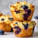 Two fluffy blueberry cottage cheese muffins stacked on a white surface with fresh blueberries scattered around.