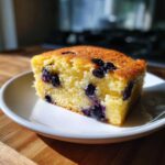 A square slice of golden, fluffy sheet pan pancakes studded with blueberries, served on a white plate.