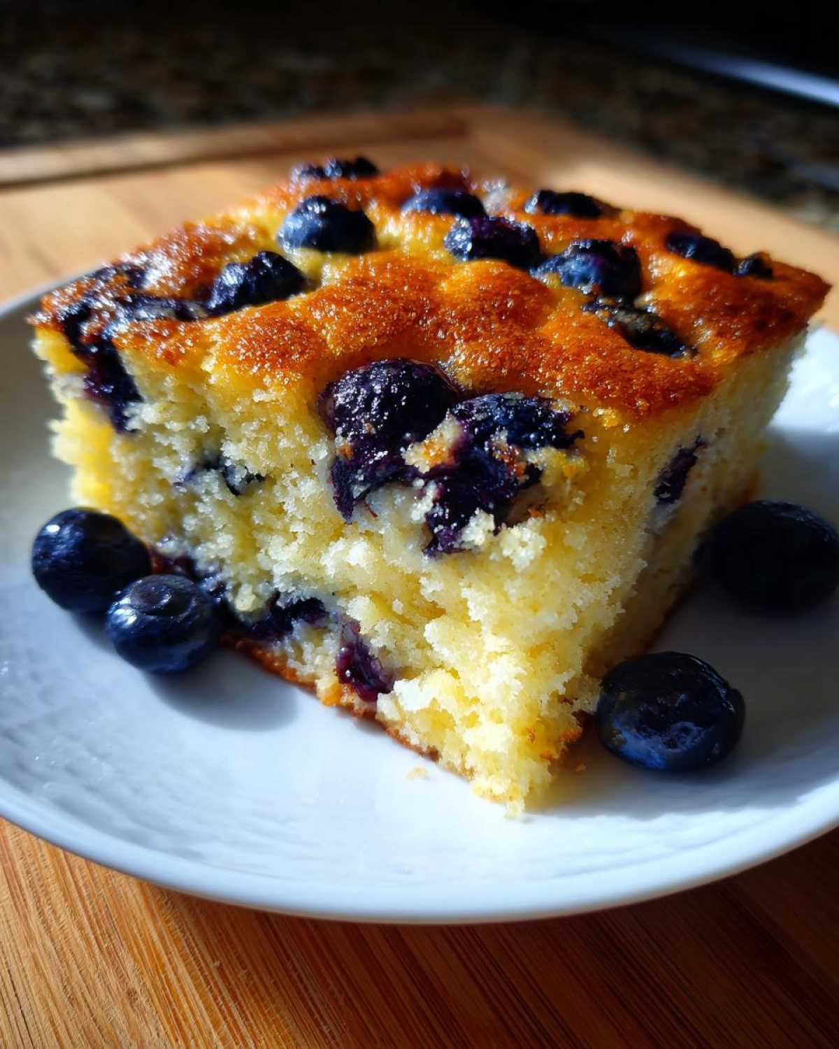 A close-up of a square slice of golden sheet pan pancakes topped with baked blueberries, served on a white plate.