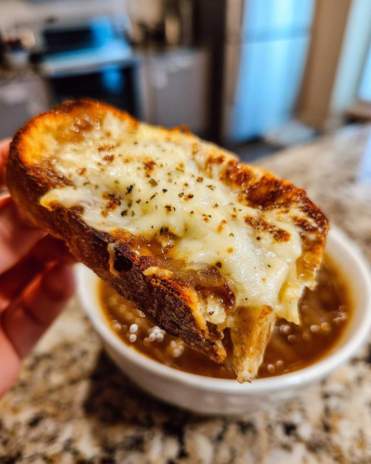A hand holding a slice of toasted bread topped with melted cheese and herbs, dipping into a bowl of crockpot french onion soup.