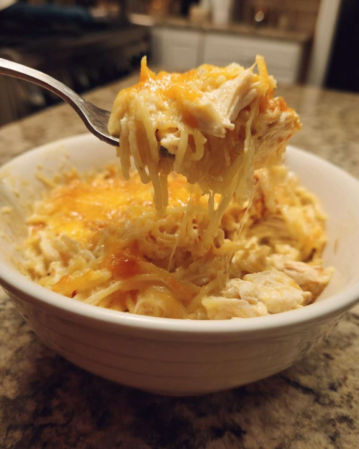 A fork lifts a cheesy, creamy bite of chicken spaghetti out of a white bowl.