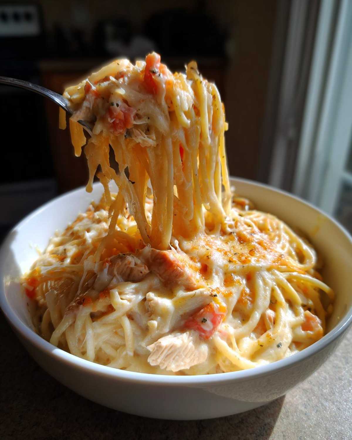 A fork lifts a generous serving of creamy, cheesy chicken spaghetti from a white bowl, showing melted cheese strings.