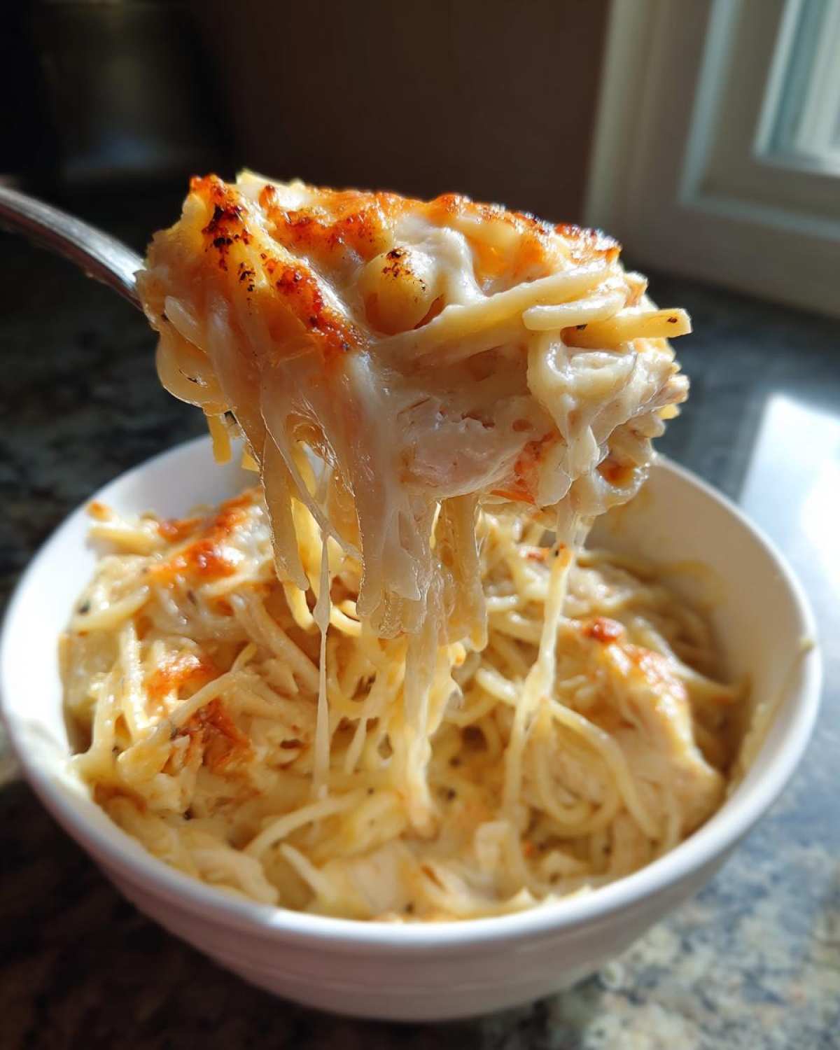 A fork lifts a cheesy, gooey portion of baked chicken spaghetti out of a white bowl.