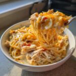 A fork lifts a cheesy, stringy portion of creamy chicken spaghetti out of a white bowl.