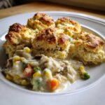 A close-up of a serving of creamy chicken pot pie with biscuits, featuring mixed vegetables like carrots and peas.