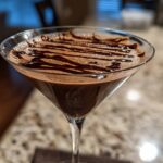 Close-up of a rich, frothy chocolate martini in a stemmed glass, topped with a dark chocolate syrup drizzle.