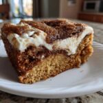 Close-up of a thick slice of cinnamon roll cake featuring a rich brown cake base, a dark cinnamon swirl, and a thick layer of white cream cheese frosting.