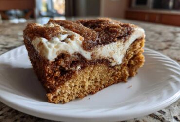 Close-up of a thick slice of cinnamon roll cake featuring a rich brown cake base, a dark cinnamon swirl, and a thick layer of white cream cheese frosting.
