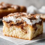Close-up of a moist slice of cinnamon roll cake featuring a swirl of cinnamon filling and topped with whipped cream and cinnamon dust.