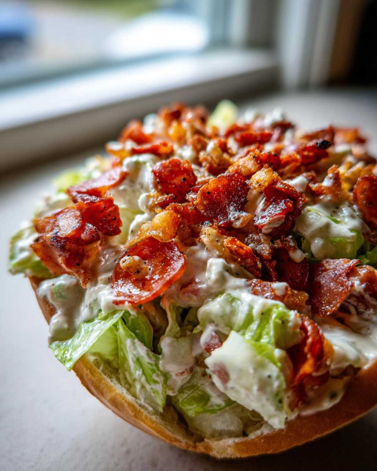 Close-up of a delicious grinder salad overflowing from a bread roll, topped with creamy dressing and crispy bacon pieces.