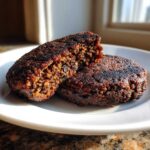 Two dark, well-seared black bean burger patties on a white plate, one broken open to show the dense, textured interior.