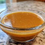 Close-up of rich, amber Copycat Apple Cider Vinaigrette dressing in a small clear glass bowl.