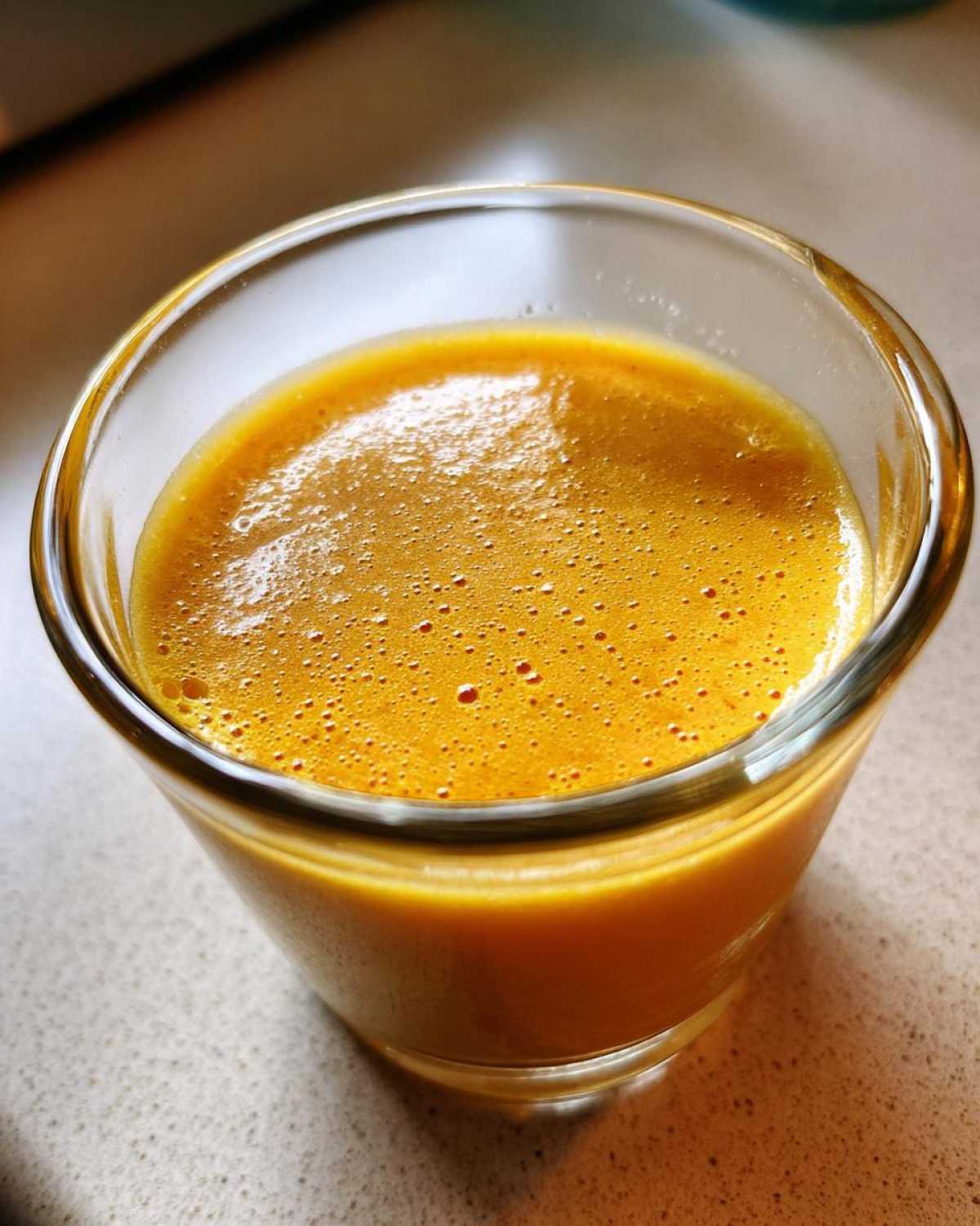 Close-up of bright orange Copycat Apple Cider Vinaigrette, slightly frothy, served in a small clear glass.