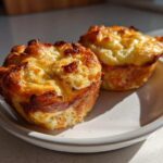 Two golden-brown Copycat Café-Style Bacon Gruyere Egg Bites served on a small white plate in bright sunlight.