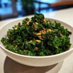 A white bowl filled with vibrant green Copycat Kale Crunch Salad topped with pine nuts and spices.
