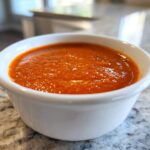 Close-up of bright orange Copycat Sweet-Tangy Polynesian Dipping Sauce served in a small white ramekin on a granite counter.