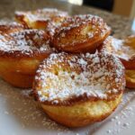 A pile of golden-brown Cozy Morning Mini German Pancakes dusted heavily with powdered sugar on a white plate.