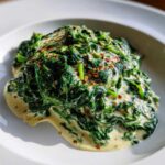 A close-up of rich, dark green creamed spinach topped with red pepper flakes served on a white plate.