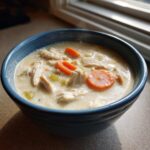 Close-up of a bowl of creamy chicken and rice soup featuring shredded chicken, sliced carrots, and celery.