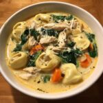 A close-up of a white bowl filled with creamy chicken tortellini soup, featuring tortellini, shredded chicken, spinach, and carrots.
