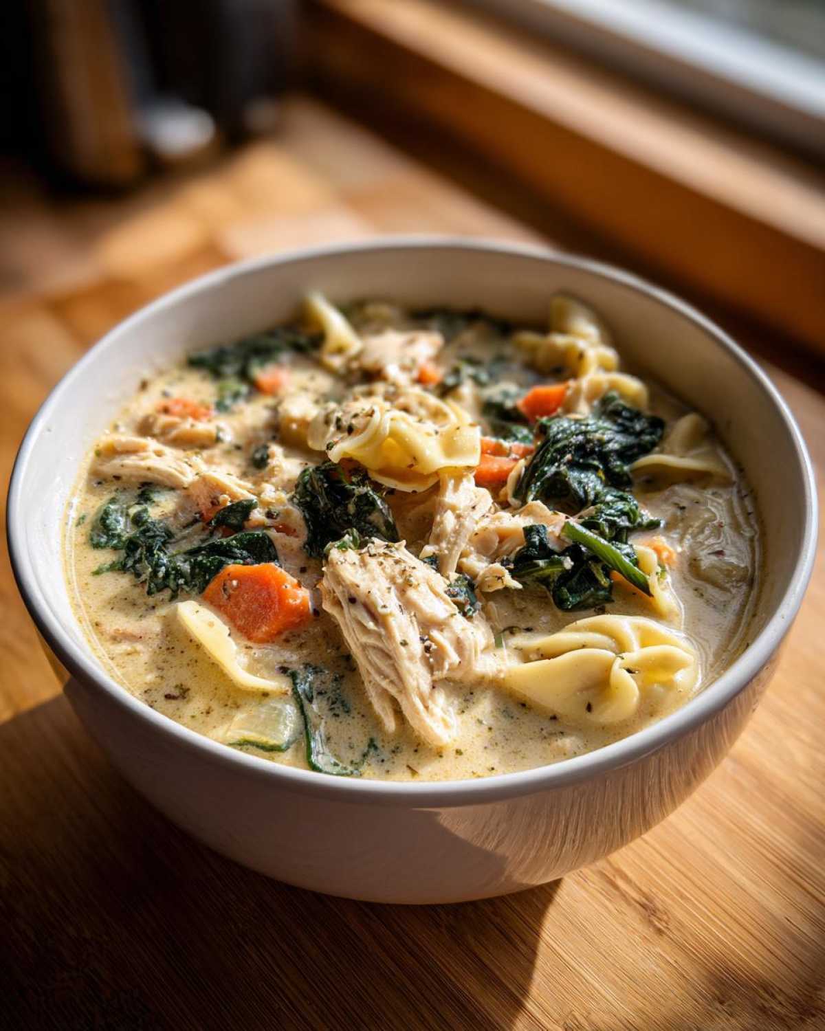A close-up of a hearty bowl of creamy chicken tortellini soup featuring shredded chicken, spinach, and carrots.