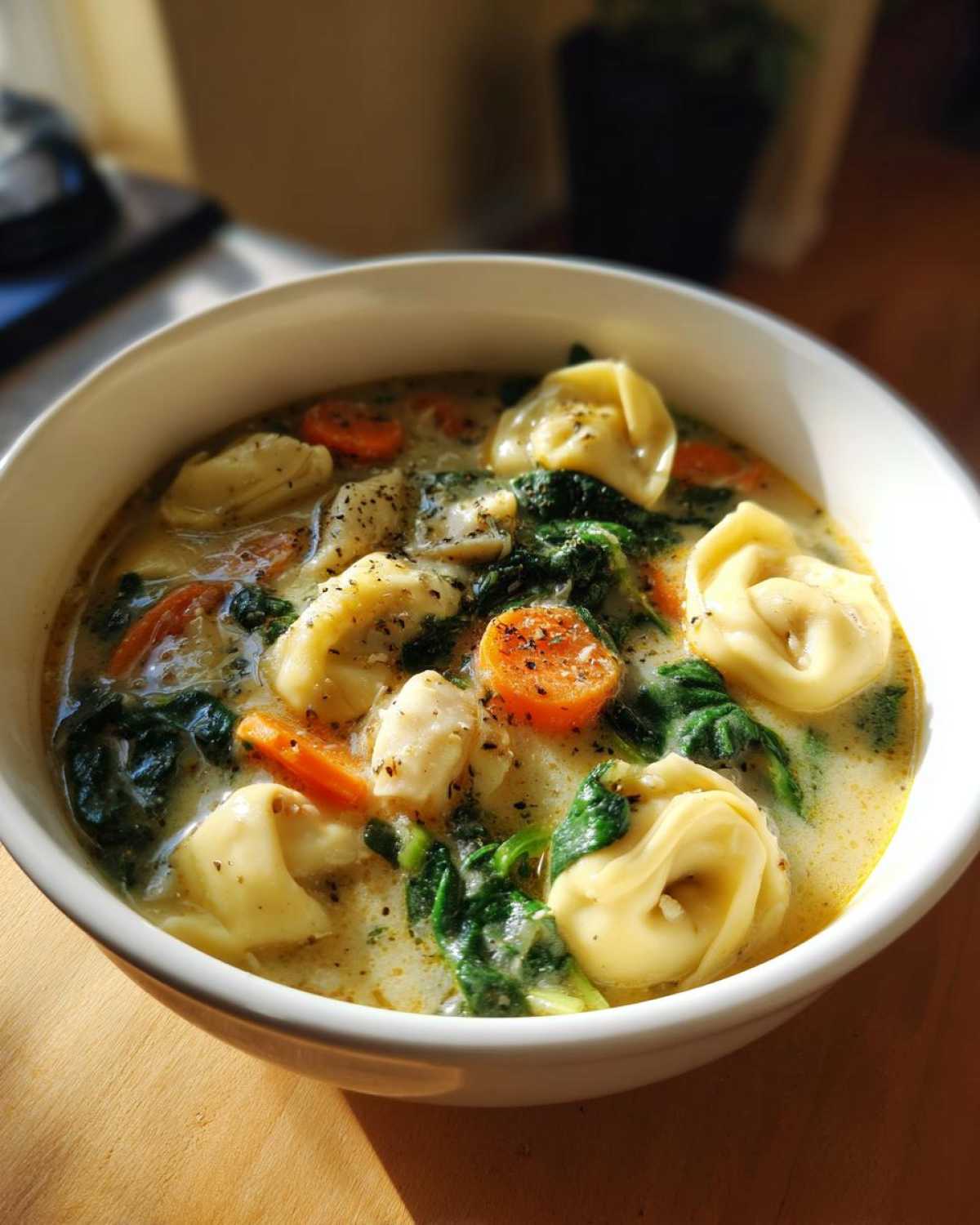 A close-up of a white bowl filled with creamy chicken tortellini soup, featuring tortellini, sliced carrots, and spinach.