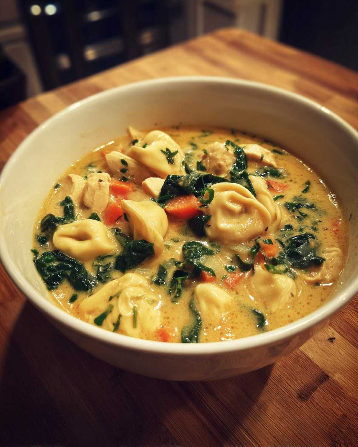 A close-up of a white bowl filled with creamy chicken tortellini soup, featuring tortellini, chicken chunks, carrots, and spinach.