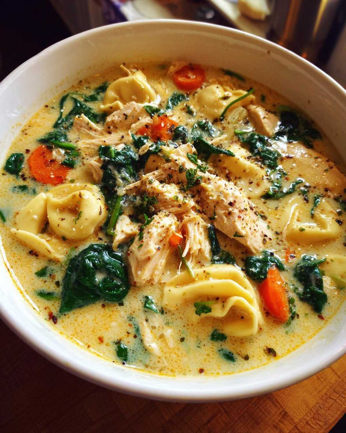 A close-up of a white bowl filled with creamy chicken tortellini soup, featuring shredded chicken, spinach, and carrots.