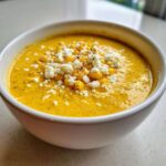 Close-up of a bowl of bright yellow creamy corn soup topped with crumbled white cheese and whole corn kernels.
