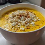 Close-up of a bowl of vibrant yellow, creamy corn soup topped generously with white crumbled cheese and spices.