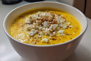 Close-up of a bowl of vibrant yellow, creamy corn soup topped generously with white crumbled cheese and spices.