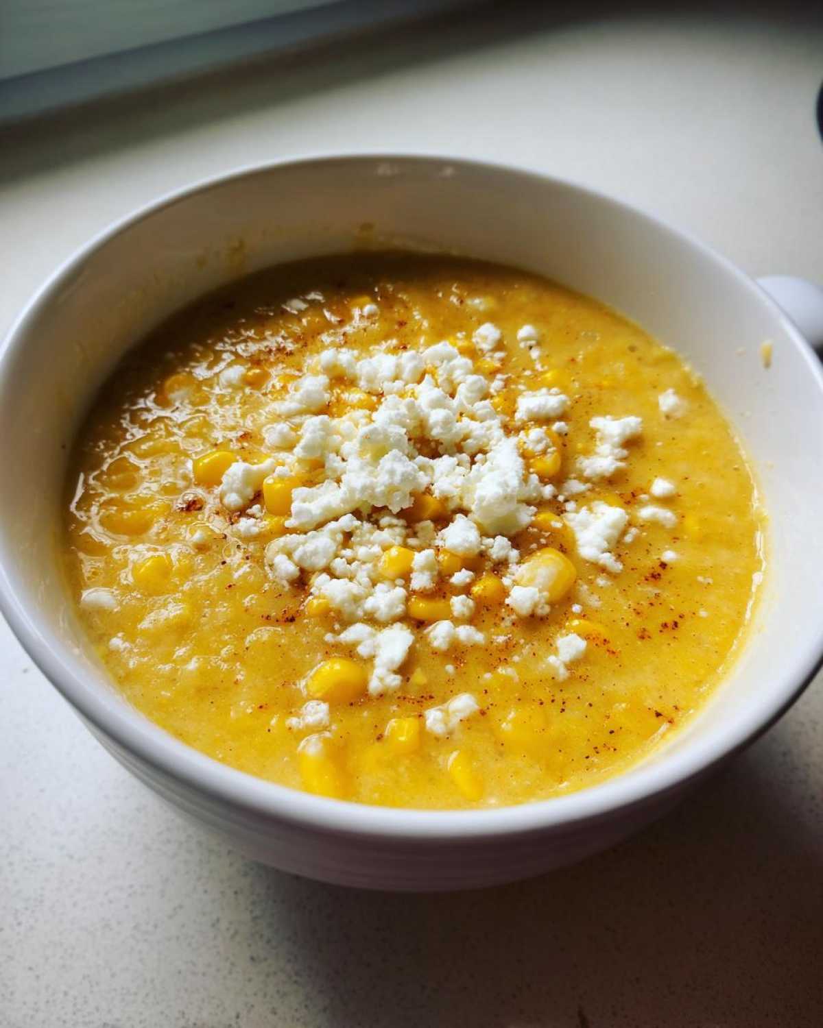 A close-up of a bowl of creamy corn soup, featuring whole kernels and topped generously with white crumbled cheese.