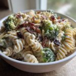 A close-up of a white bowl filled with Creamy Pasta Salad with Bacon and Broccoli, featuring rotini pasta and a rich white dressing.