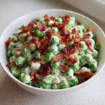 A white bowl filled with bright green peas coated in a creamy dressing and topped generously with crispy bacon bits, showcasing the Creamy Pea Salad.