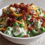 A white bowl filled with bright green peas coated in a creamy dressing, topped with crumbled bacon and shredded cheddar cheese, showcasing the Creamy Pea Salad.