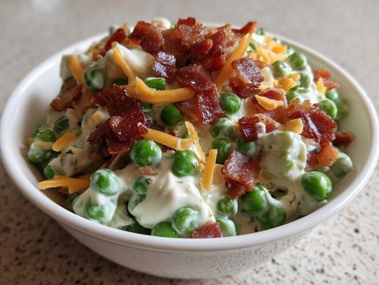 A white bowl filled with bright green peas coated in a creamy dressing, topped with crumbled bacon and shredded cheddar cheese, showcasing the Creamy Pea Salad.