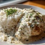 A close-up of a serving of Creamy Smothered Chicken and Rice, featuring a piece of chicken covered in rich white sauce next to fluffy white rice.