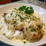 A close-up of Creamy Smothered Chicken and Rice, featuring a chicken breast covered in rich sauce over white rice, garnished with parsley.