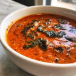 Close-up of creamy, vibrant orange tomato basil soup topped with wilted basil leaves in a white bowl.